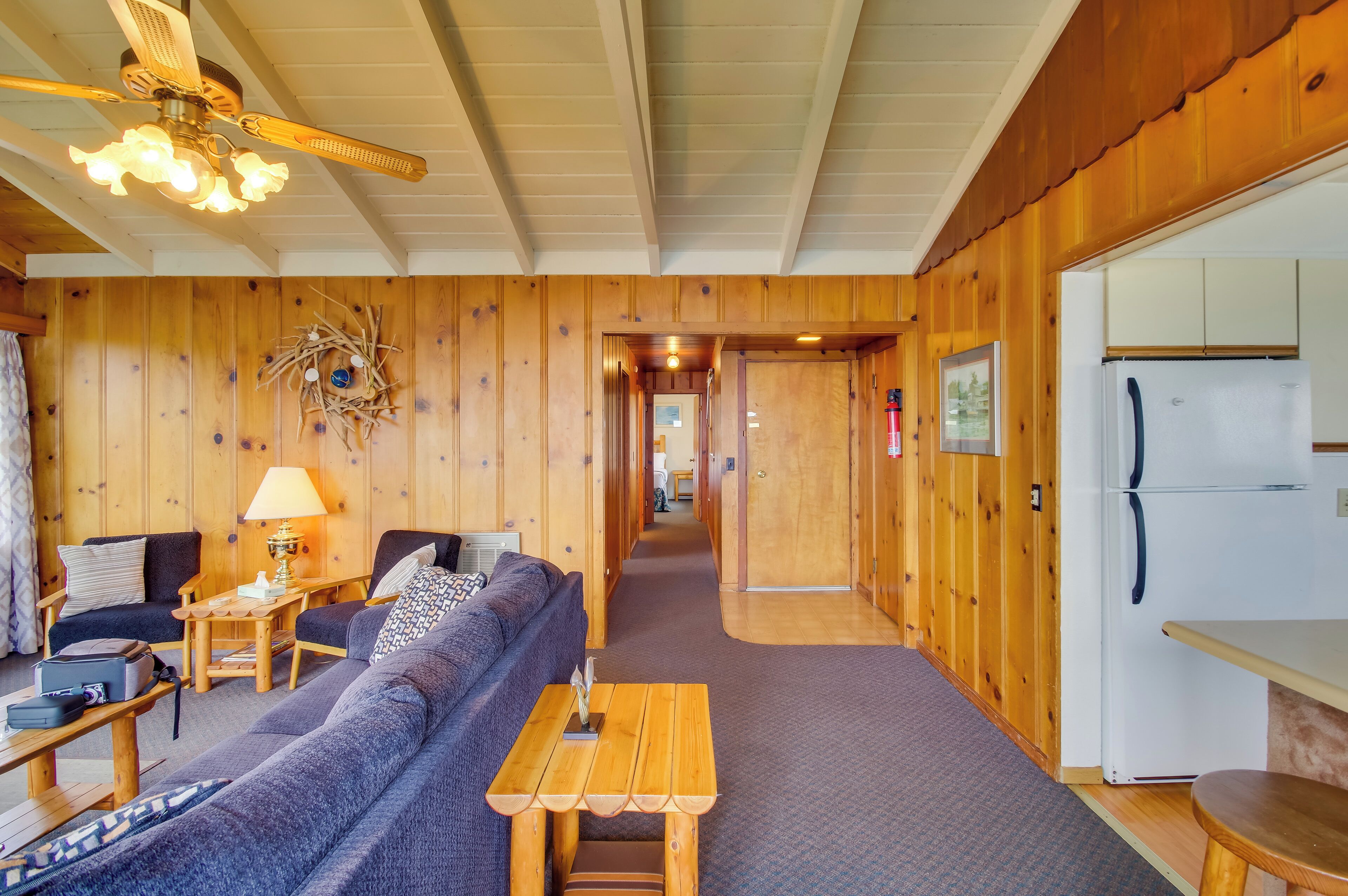 A cozy cabin interior with a living area, wooden walls, a ceiling fan, a refrigerator, and a hallway leading to another room.
