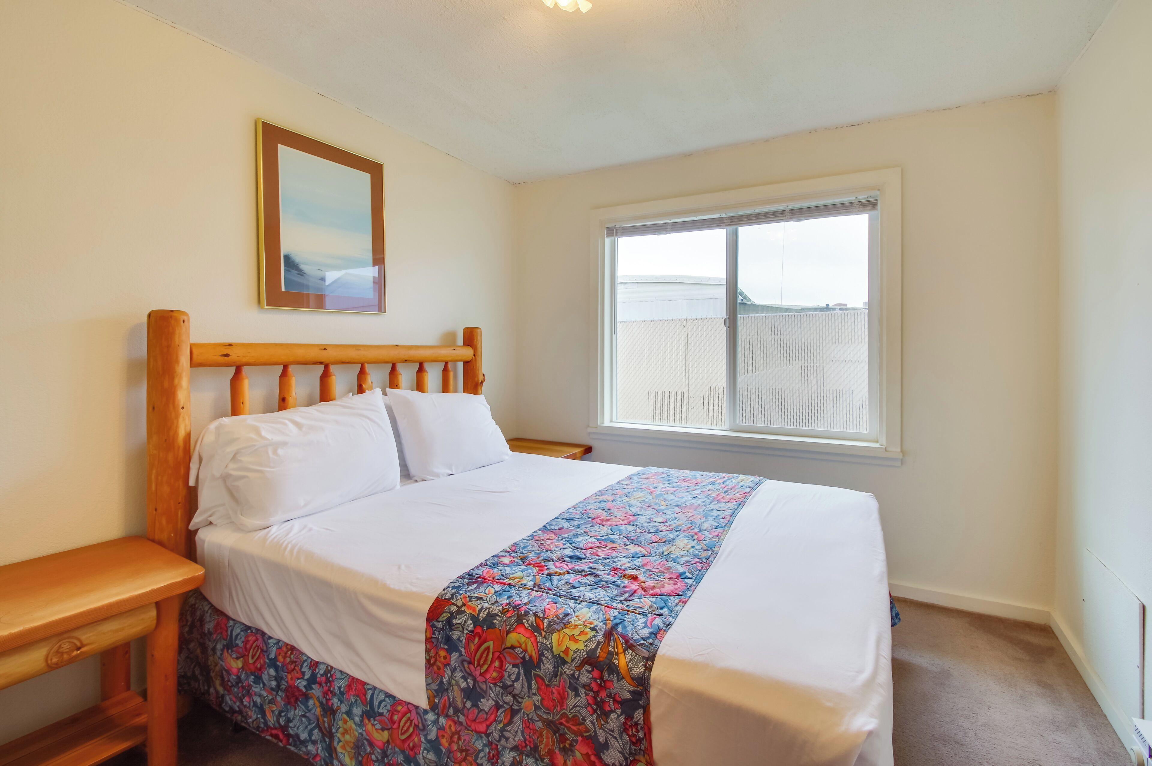 A bedroom with a wooden bed frame, white bedding, a floral bedspread, a nightstand, a window with a view, and a framed picture on the wall.