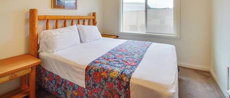 A bedroom with a wooden bed frame, white bedding, a floral bedspread, a nightstand, a window with a view, and a framed picture on the wall.