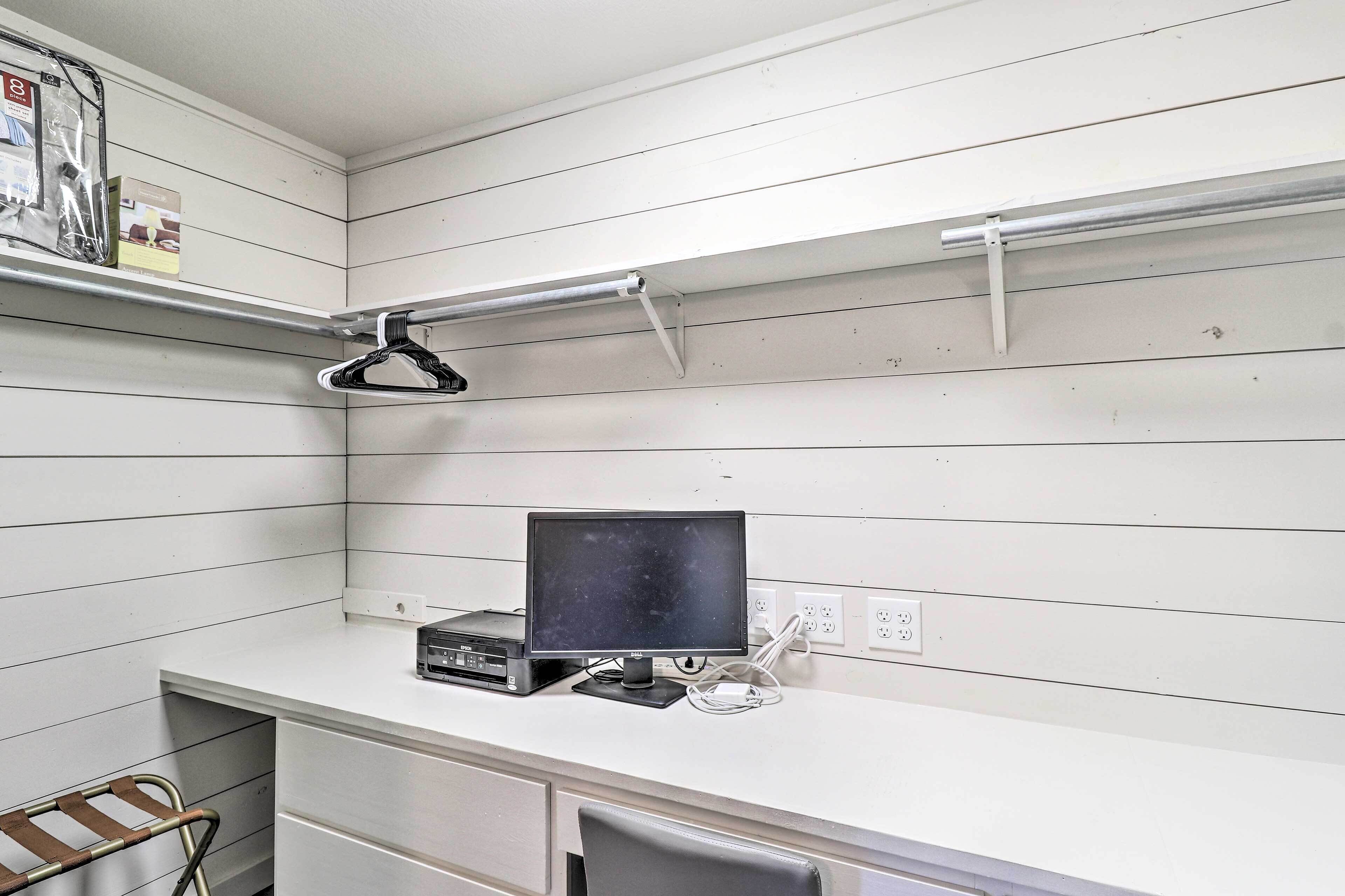 A small office space with a desk, a computer monitor, and a chair. There are shelves above the desk and a hanging light fixture.