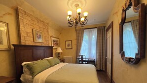 Cama bem arrumada com lençóis verdes e brancos, cabeceira de madeira, candelabro, quadro emoldurado acima da cama, uma cadeira perto da janela e um espelho grande na parede.