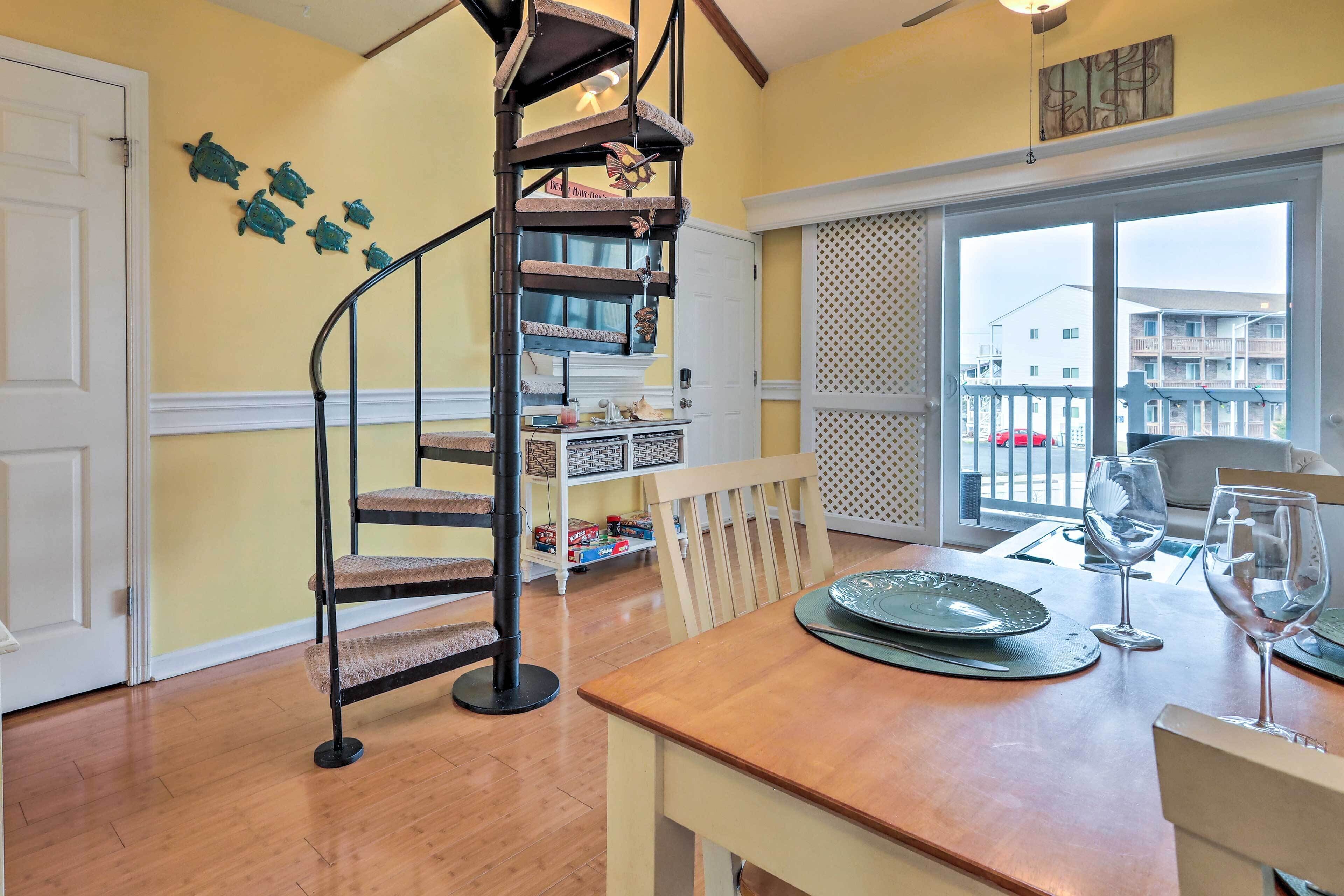 A dining area with a wooden table set for two, a spiral staircase, and a view of the outside balcony.
