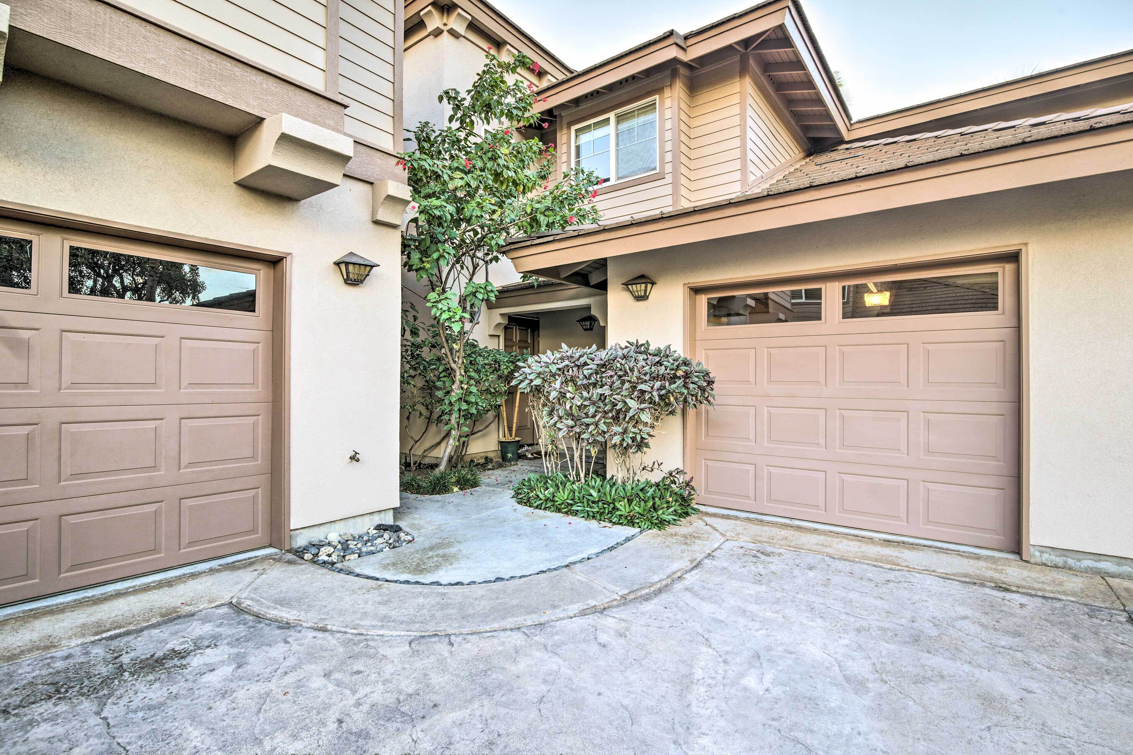 A residential driveway with two garage doors, a concrete pathway, and a small garden area with shrubs and a tree.