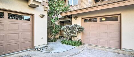 A residential driveway with two garage doors, a concrete pathway, and a small garden area with shrubs and a tree.