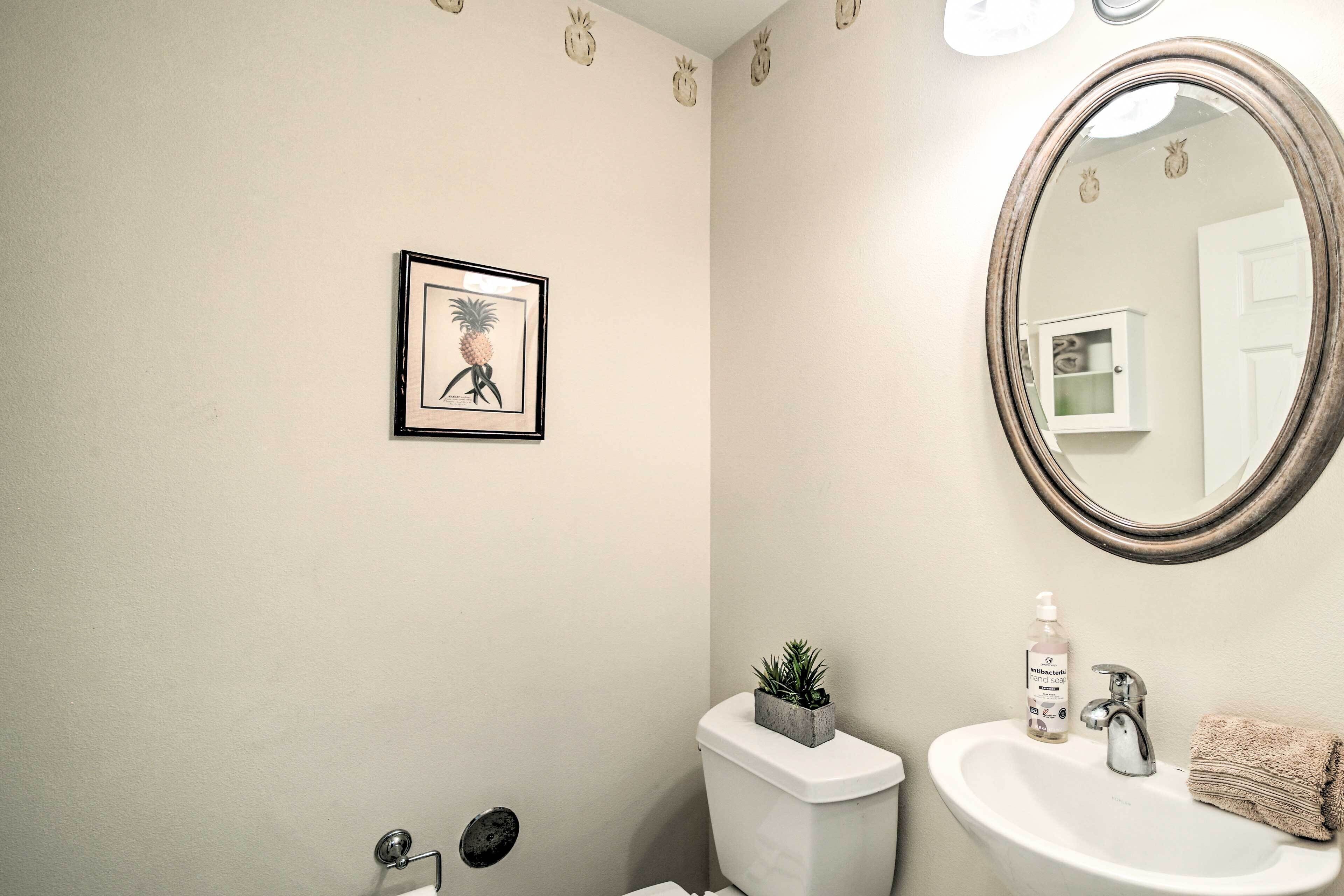 Ein Badezimmer mit Toilette, Waschbecken und Spiegel. An der Wand hängt ein gerahmtes Bild und auf dem Toilettenbecken steht eine kleine Pflanze.
