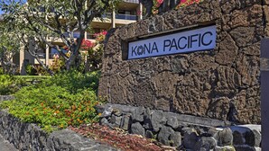 A stone wall with a sign reading "KONA PACIFIC" and a landscaped area with plants and flowers.