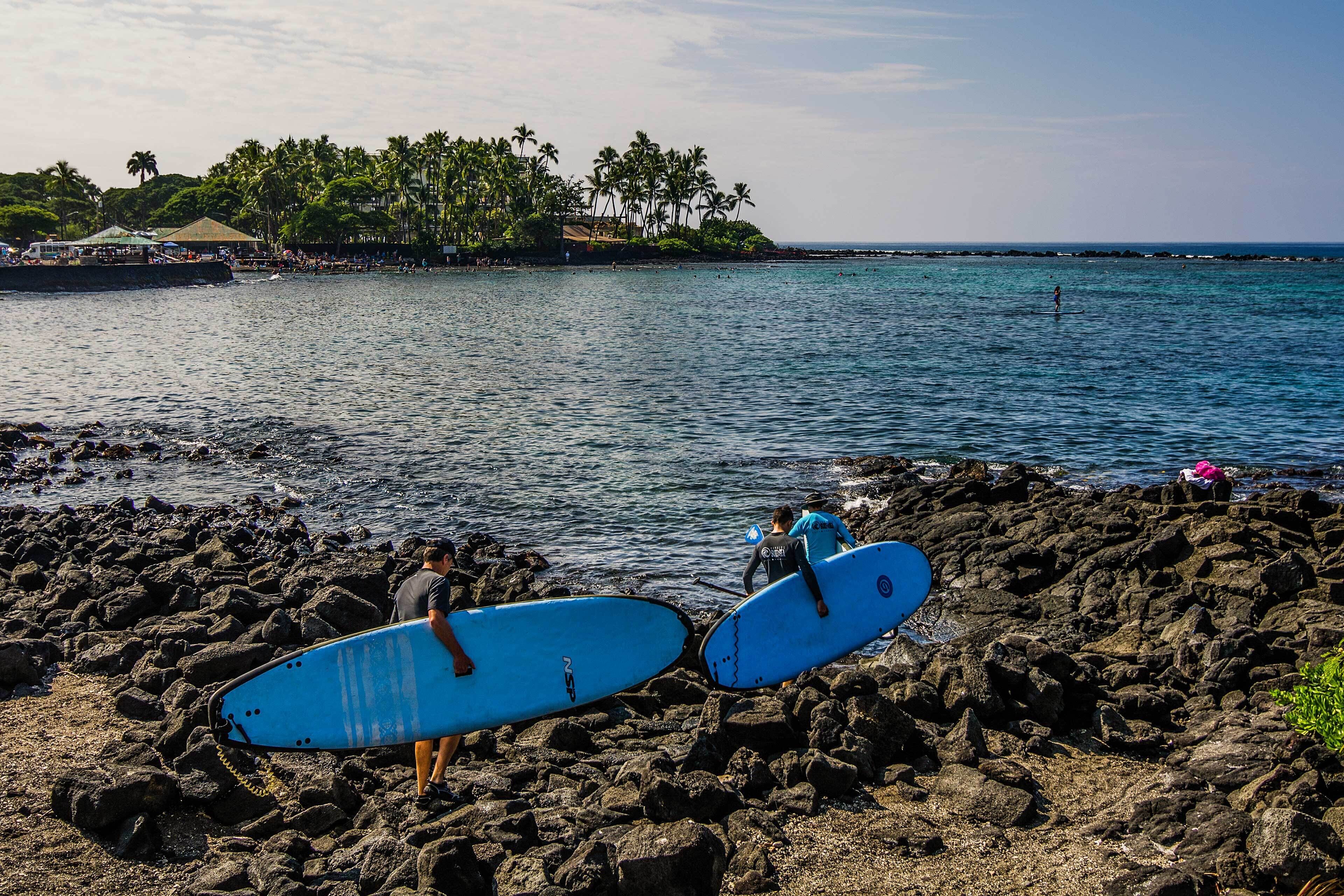 A rocky beach with surfers carrying surfboards, a clear sky, and a distant island with palm trees.