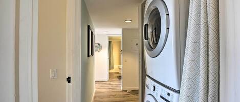 Una lavandería con dos lavadoras y secadoras apiladas, piso de madera y una puerta que da a otra habitación.