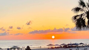 Ein Strandbild mit Sonnenuntergang, Reetdachschirmen, Liegestühlen und Blick auf den Ozean.