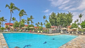 A swimming pool with clear blue water, surrounded by lounge chairs and umbrellas, with palm trees and buildings in the background. - Ultimate Oceanfront Townhome w/ A/C On Kona Coast (Kailua-Kona)