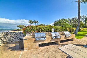 An outdoor barbecue area with stone countertops and grills, overlooking a landscaped garden and the ocean.
