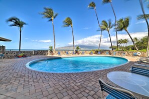 A swimming pool with a stone wall and palm trees, surrounded by lounge chairs and a table.