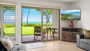 Een woonkamer met een schuifdeur die leidt naar een balkon met uitzicht op de oceaan. Er staat een houten tafel met stoelen, een aan de muur bevestigde televisie en een kast met decoratieve objecten.