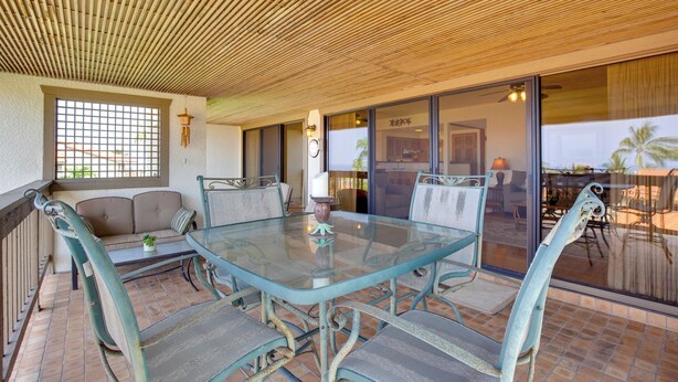 A covered patio area with a glass table and metal chairs, a cushioned bench, and a view through a glass door to an outdoor patio. - Kailua-kona Condo w/ Resort Access & Ocean View! (Kailua-Kona)