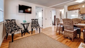 A modern living room with a flat-screen TV, two patterned chairs, a small table, and a dining area with wicker chairs.