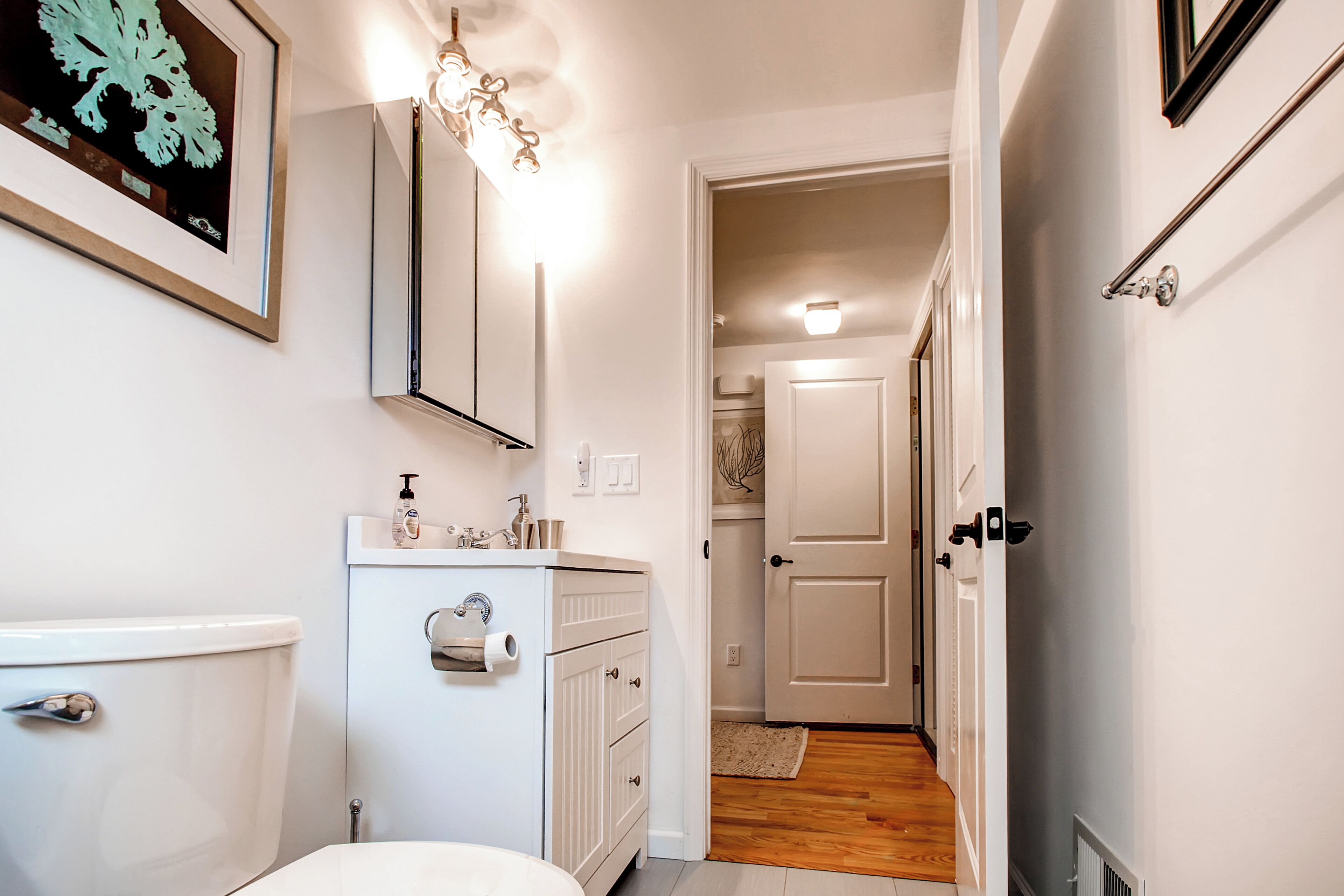 A bathroom with a toilet, sink, and cabinet. There is a framed artwork on the wall and a light fixture above the sink.