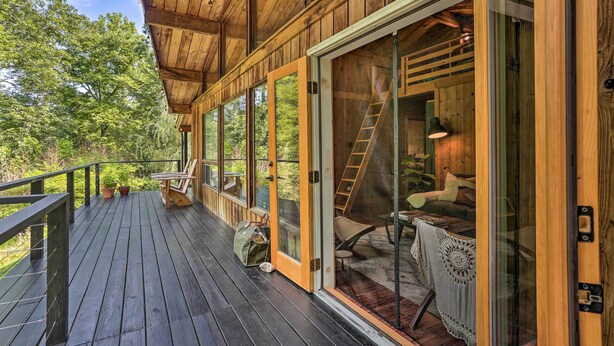 A wooden deck with a glass door leading to a cozy interior with a staircase, seating, and a table. - High Falls Restorative Cabin in the Woods! (High Falls)