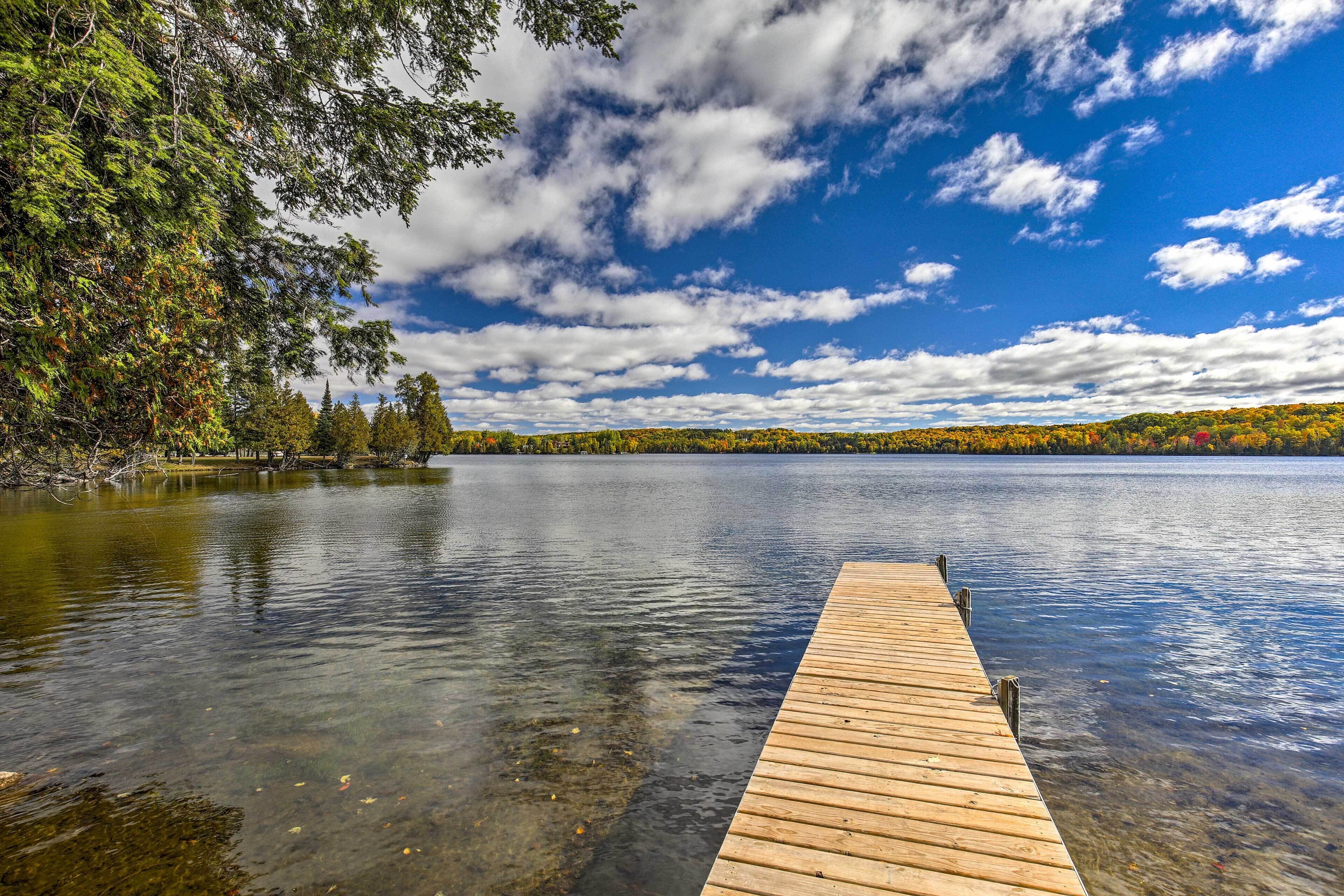 Un muelle de madera se extiende sobre un lago tranquilo con aguas cristalinas, rodeado de árboles y un cielo parcialmente nublado.