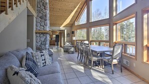 A spacious living area with a stone fireplace, a dining table with chairs, and large windows offering a view of the outdoors. - Secluded Lakehouse w/ Private Dock + Serene Views! (Iron River)
