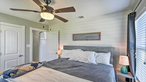 Une chambre à coucher avec un lit, un ventilateur de plafond, une table de chevet avec une lampe, une fenêtre avec des stores et un tableau accroché au mur.