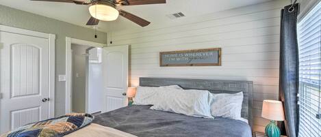 A bedroom with a bed, a ceiling fan, a nightstand with a lamp, a window with blinds, and a sign on the wall.