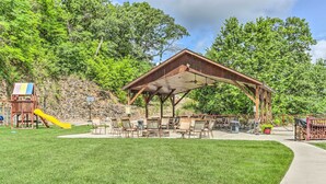 Ein überdachter Pavillon mit Gartenmöbeln, ein Spielplatz mit einer gelben Rutsche und ein bewaldeter Hang im Hintergrund.
