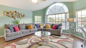 A living room with a sectional sofa, a glass coffee table, a ceiling fan, and large windows.