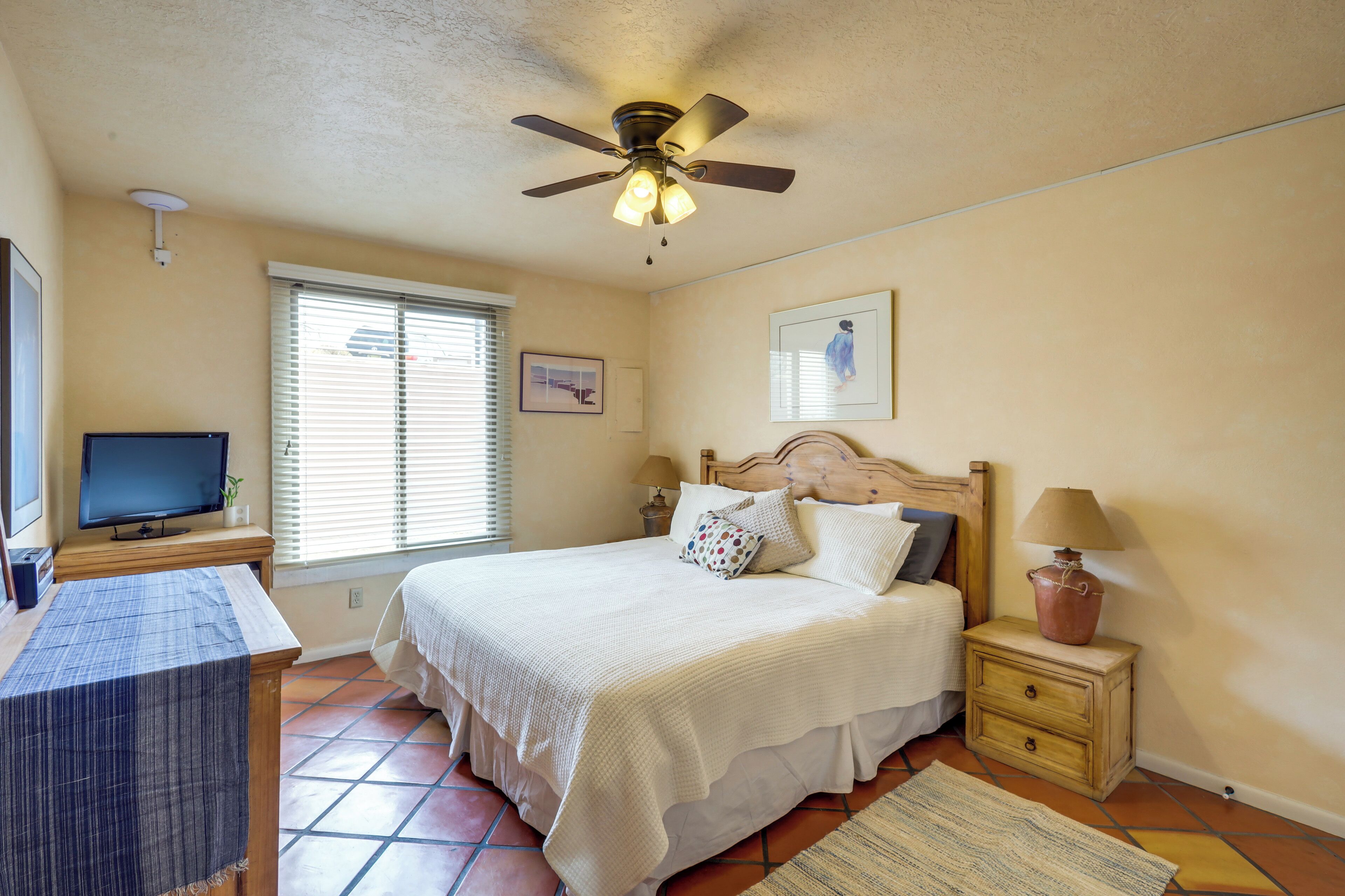 A bedroom with a bed, bedside tables, a ceiling fan, a TV, and a window with blinds.