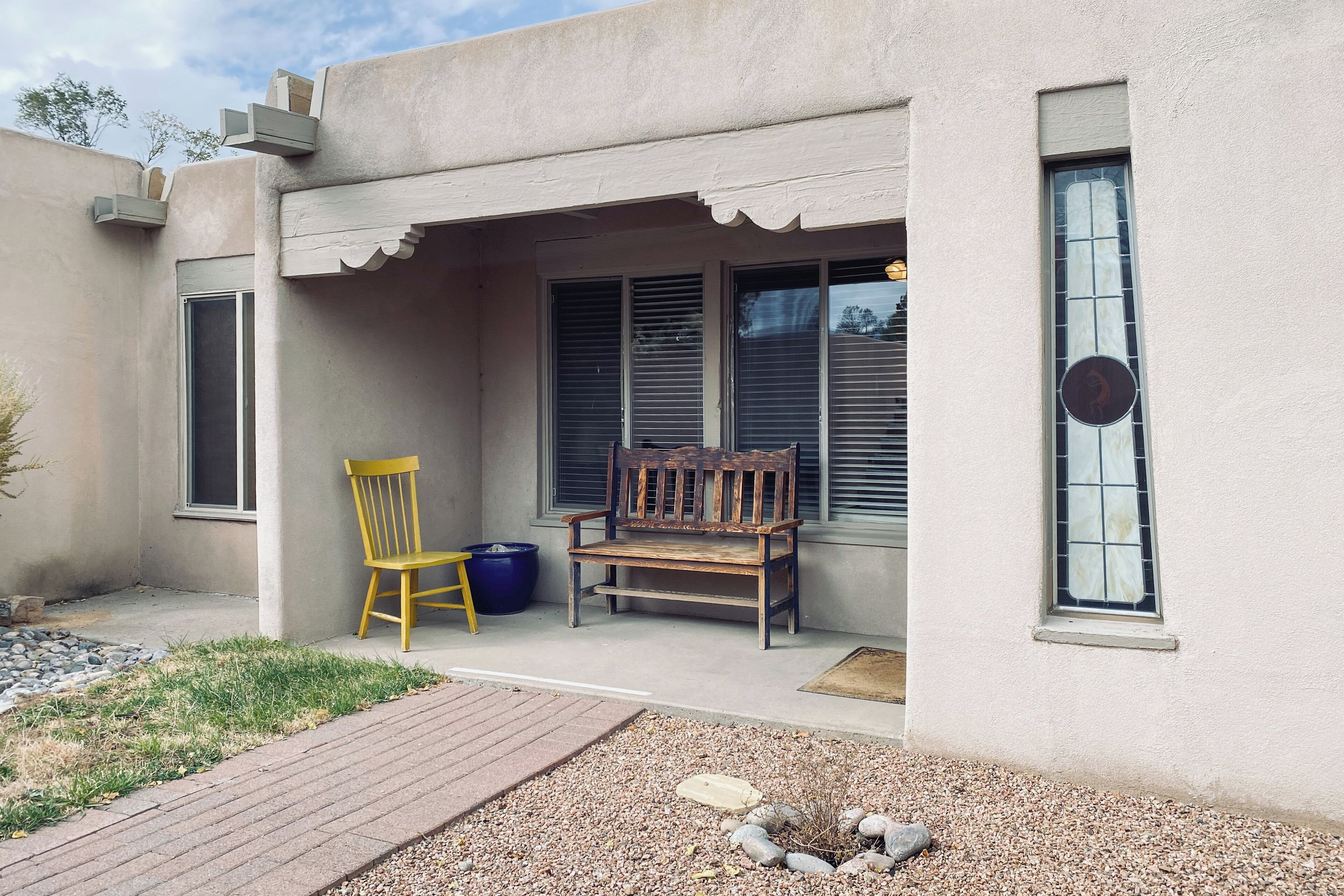 A building with a covered patio area, a wooden bench, a yellow chair, and a blue bucket.