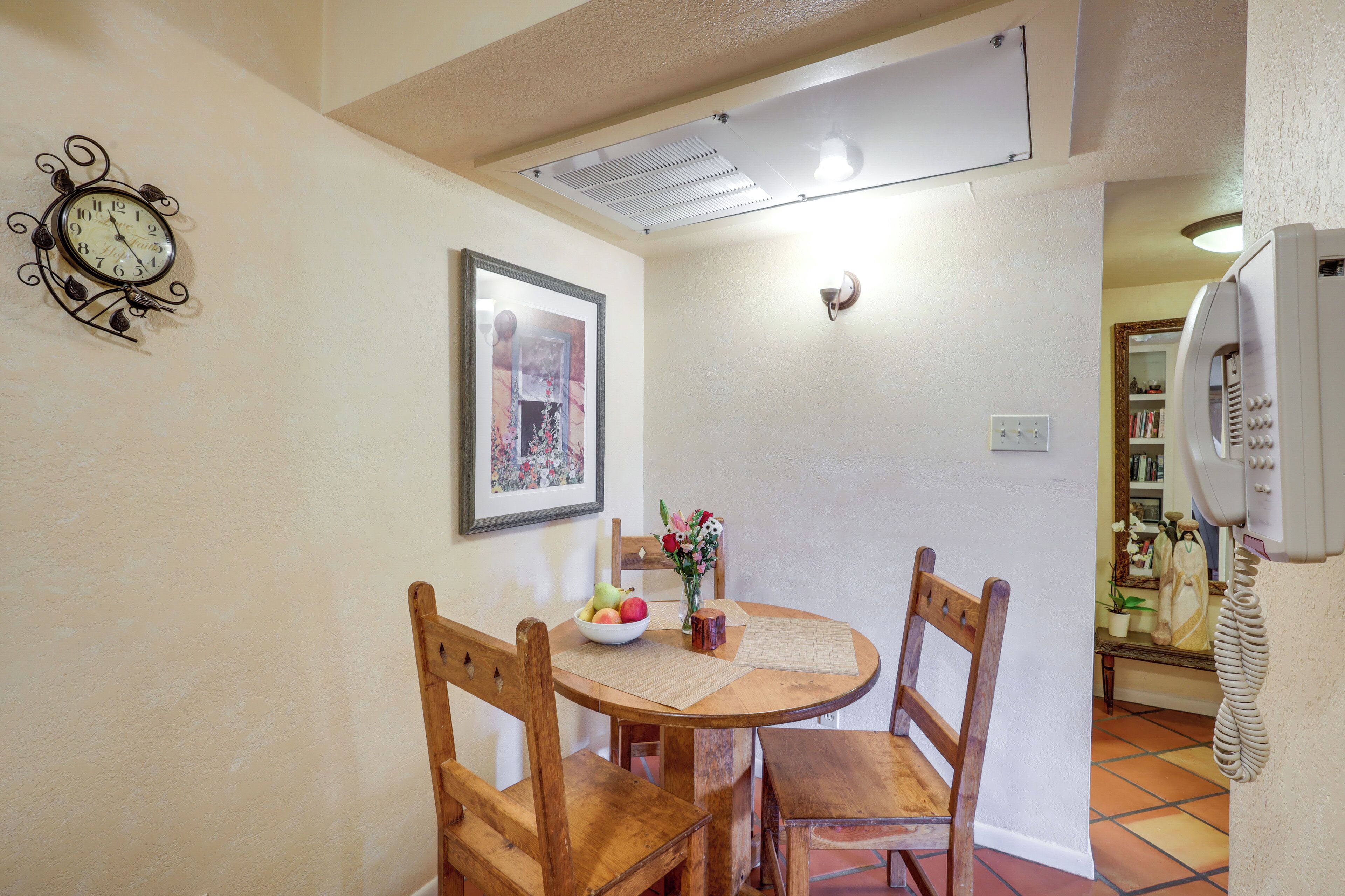 A dining area with a round wooden table, four wooden chairs, a clock, a framed picture, and a vase with flowers.
