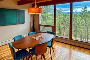 A dining area with a round table, six chairs, a hanging light fixture, and a view of a forest through large windows. - Chic Boulder Mountain Home w/ Hot Tub + Views (Boulder)