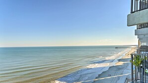 A beach view from a balcony with a clear sky, a sandy beach, and a pier in the distance. - Oceanfront Oasis w/ Deck and Resort Beach Access! (Murrells Inlet)