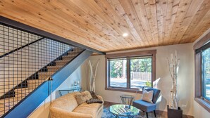Un salon moderne avec un plafond en bois, un escalier doté d’une rampe en métal, un canapé beige, un fauteuil bleu et une table basse.