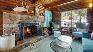 A cozy cabin interior with a stone fireplace, wooden walls, a comfortable seating area, and a large window offering a view of the outside. - Boutique + Artsy Log Cabin in North Lake Tahoe! (Kings Beach)