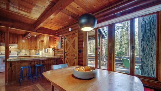 A wooden cabin interior with a kitchen, dining area, and large windows overlooking a forest. - Boutique + Artsy Log Cabin in North Lake Tahoe! (Kings Beach)