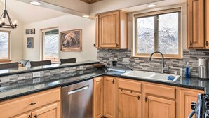 A modern kitchen with wooden cabinets, a granite countertop, and a large window providing a view of the outdoors. - Alaskan Mountain Gem With Private Hot Tub & Gym! (Eagle River)