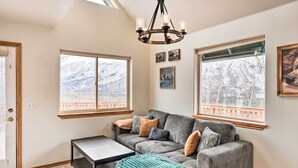A cozy living room with a sectional sofa, a coffee table, and a chandelier. There are large windows with a view of snow-covered mountains. - Alaskan Mountain Gem With Private Hot Tub & Gym! (Eagle River)