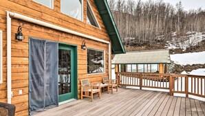 A wooden cabin with a deck, a green door, and a window overlooking a snowy landscape. - Alaskan Mountain Gem With Private Hot Tub & Gym! (Eagle River)