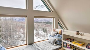 A room with a large window seat overlooking a snowy landscape with trees and mountains. There is a bookshelf with various board games and books. - Alaskan Mountain Gem With Private Hot Tub & Gym! (Eagle River)