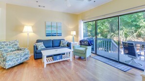 A living room with a sofa, armchairs, a coffee table, and a TV. There is a ceiling fan, a window with a view of trees, and a door leading to a balcony.