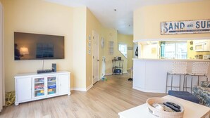 A living room with a TV, a white cabinet with glass doors, a kitchen area with a bar, and a sign that says "SAND AND SURF".