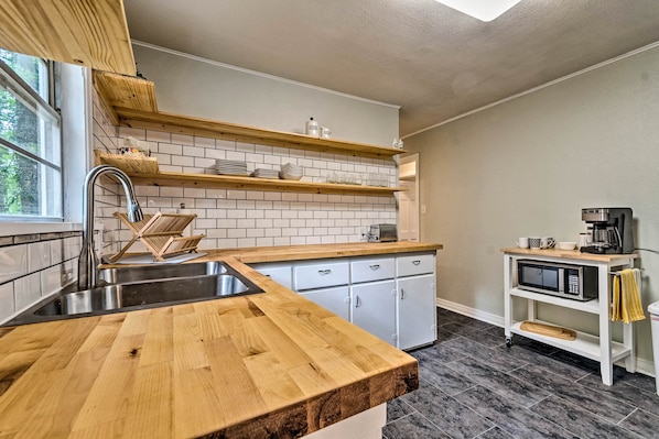 Una cocina con encimera de madera, armarios blancos, un lavamanos de acero inoxidable y un microondas sobre un estante blanco.