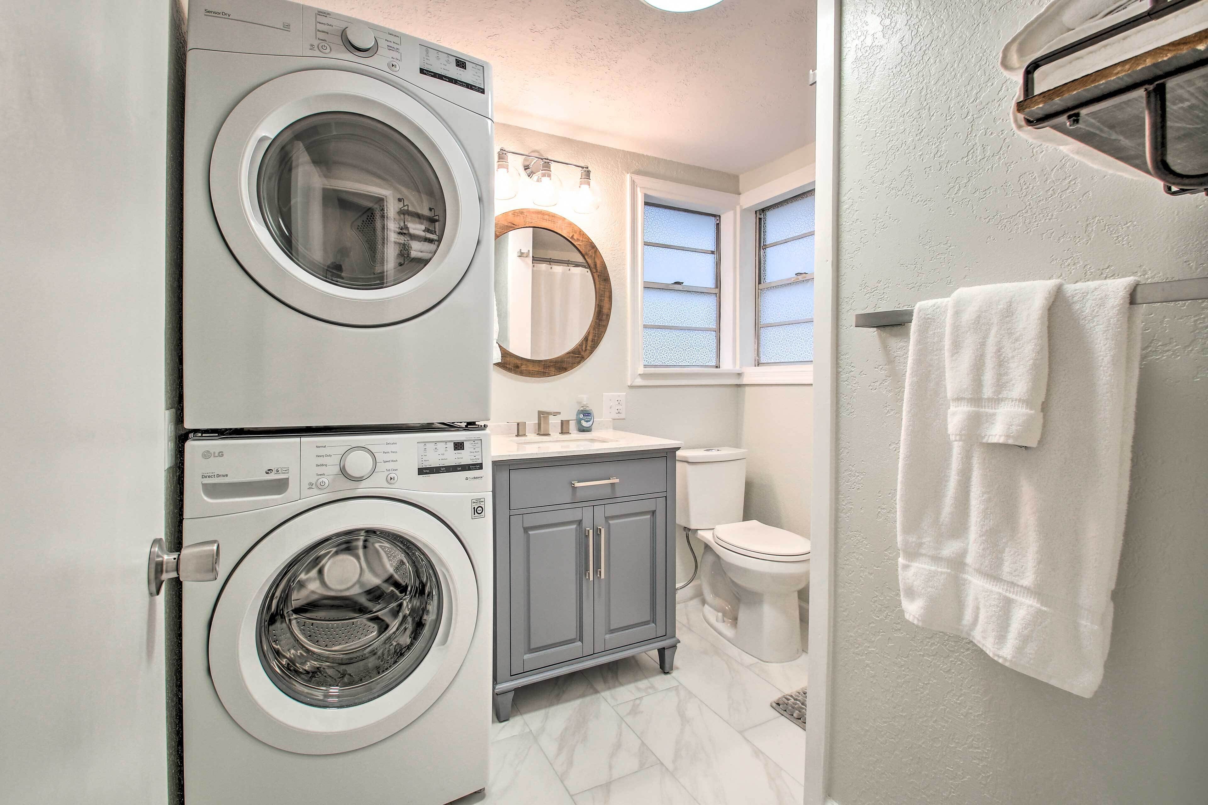 A compact laundry room with a front-loading washer and dryer, a gray cabinet, a toilet, and a window with a towel hanging on the wall.