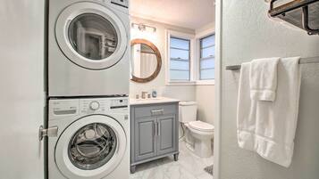 A compact laundry room with a front-loading washer and dryer, a gray cabinet, a toilet, and a window with a towel hanging on the wall.