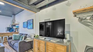 Un soggiorno moderno con un divano grigio, mobili contenitori in legno, un televisore a schermo piatto e, sullo sfondo, un angolo cottura.