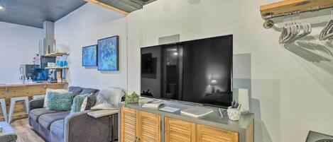 A modern living room with a grey sofa, wooden cabinets, a flat-screen TV, and a kitchen area in the background.