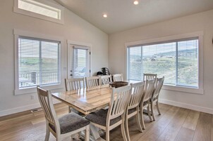 Uma sala de jantar com mesa de madeira e seis cadeiras, janelas amplas com persianas e um respiradouro no teto.