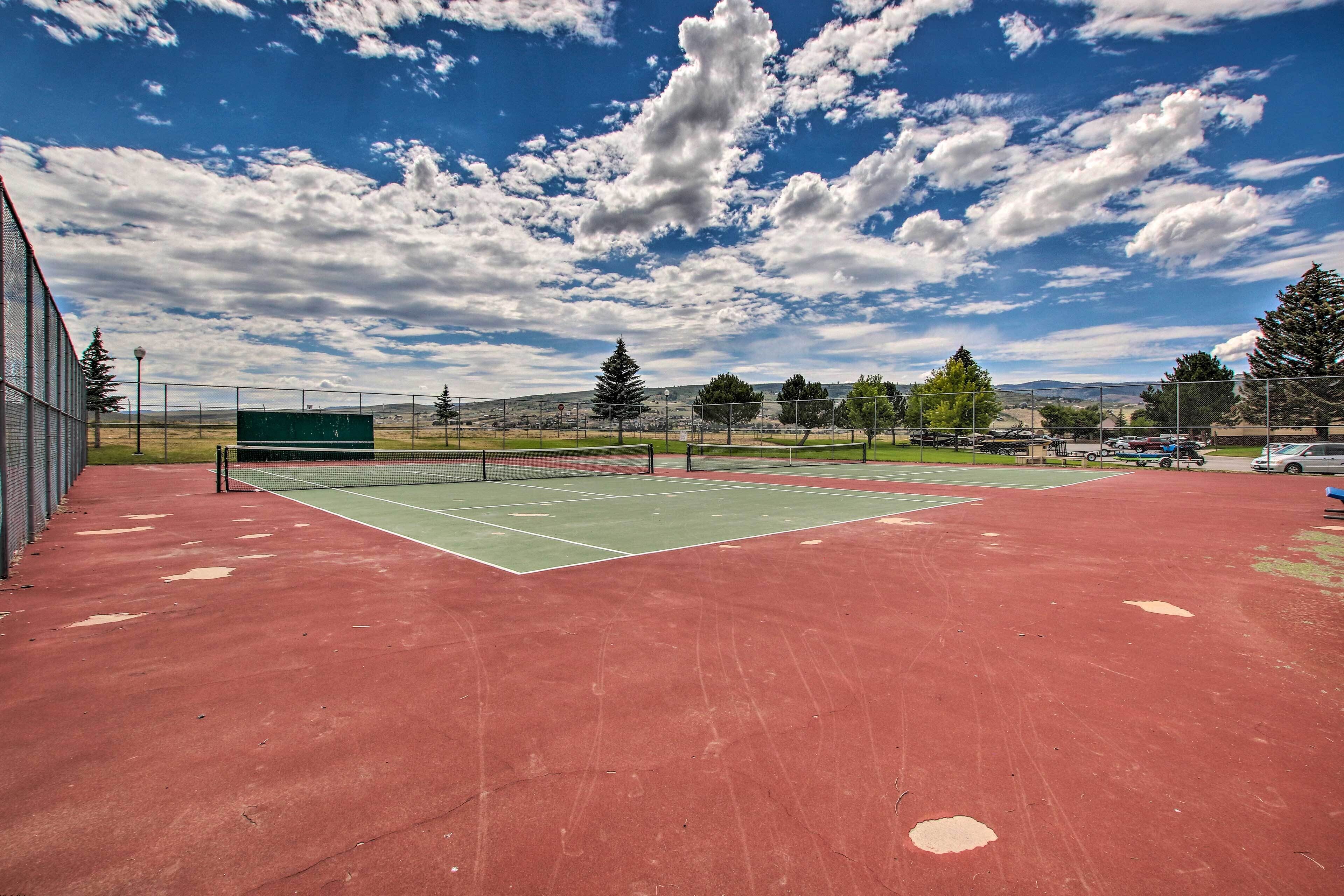 Una cancha de tenis exterior con superficie roja, rodeada de árboles y un cielo despejado.