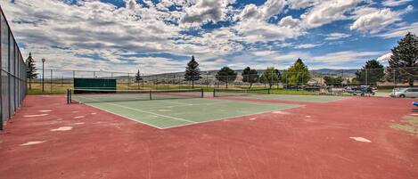 Una cancha de tenis exterior con superficie roja, rodeada de árboles y un cielo despejado.