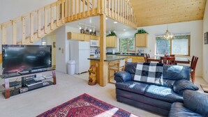 Ein geräumiger Wohnbereich mit großem Fernseher, einer Couch, einem roten Teppich mit Muster und einer Holztreppe. Im Hintergrund ist eine Küche zu sehen.